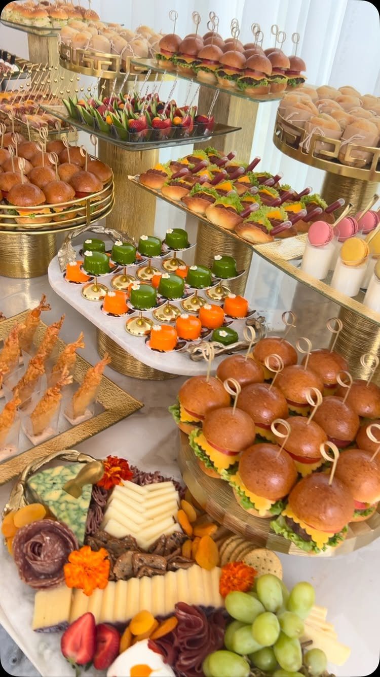 Gourmet catering spread with mini burgers, charcuterie, and appetizers on gold tiered stands.