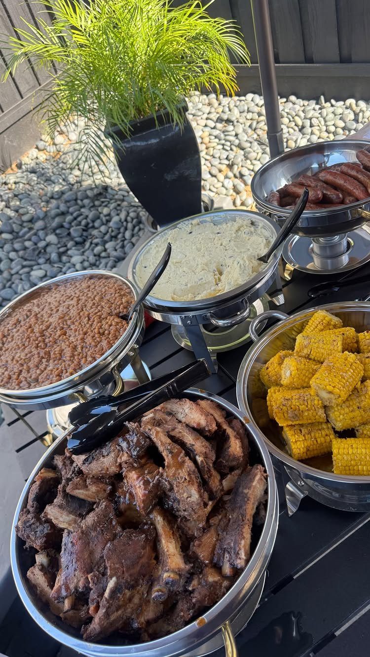 Buffet spread with ribs, corn, and beans in silver dishes outdoors.