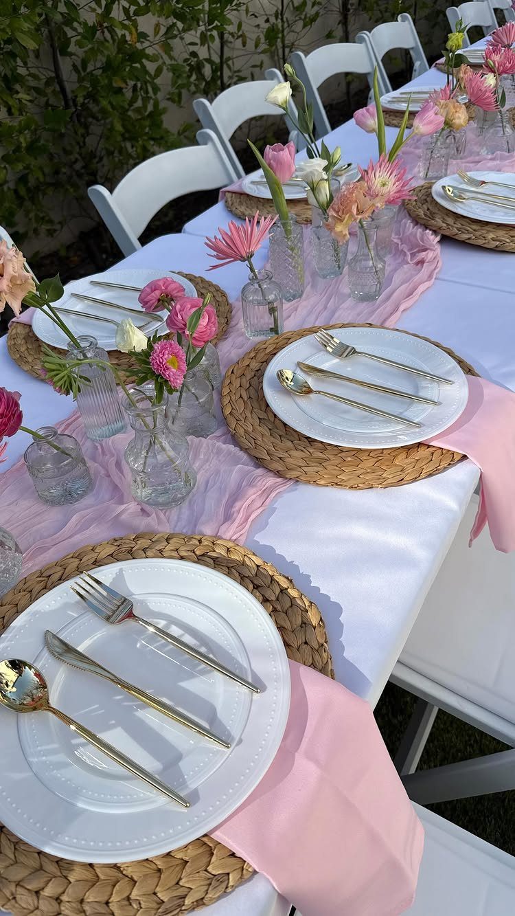 Outdoor dinner table set with white plates, gold cutlery, and pink floral arrangements.
