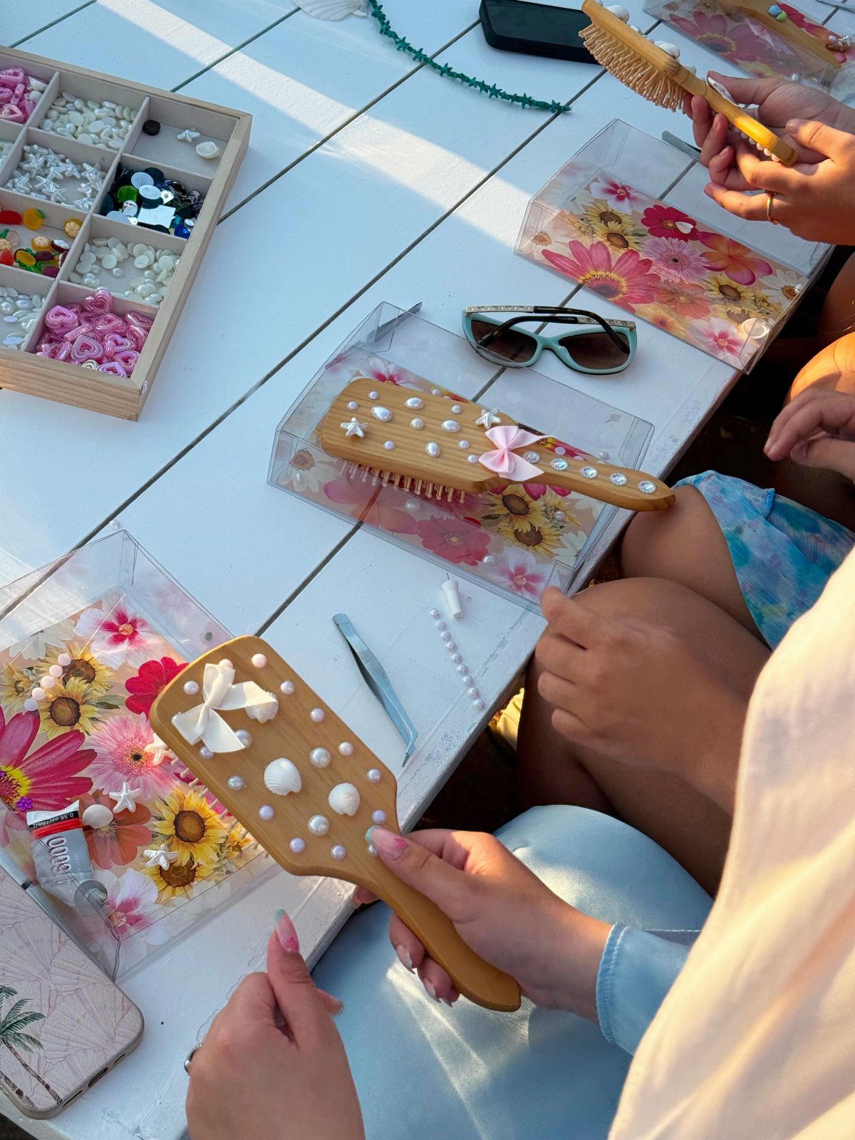 People decorating wooden hairbrushes with pearls and bows at an outdoor party table