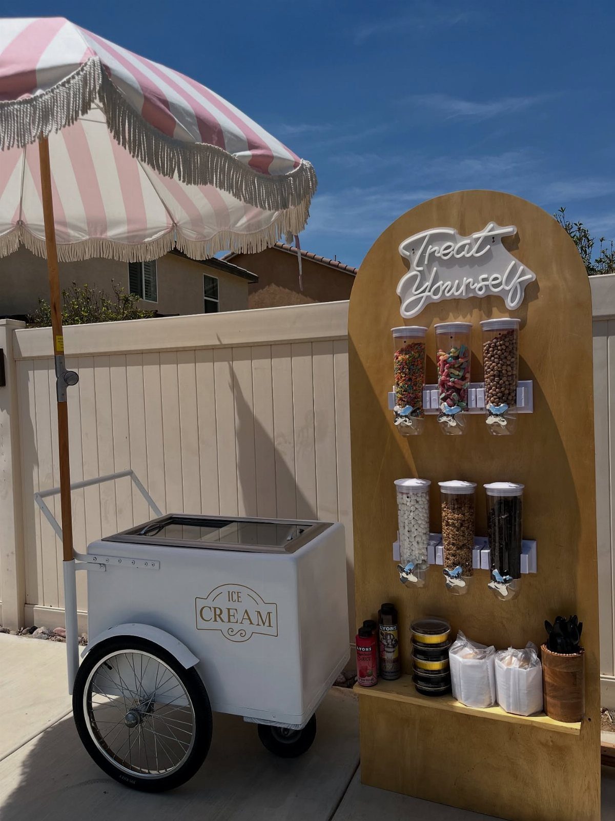 White ice cream cart and wooden topping station with candy dispensers outdoors.