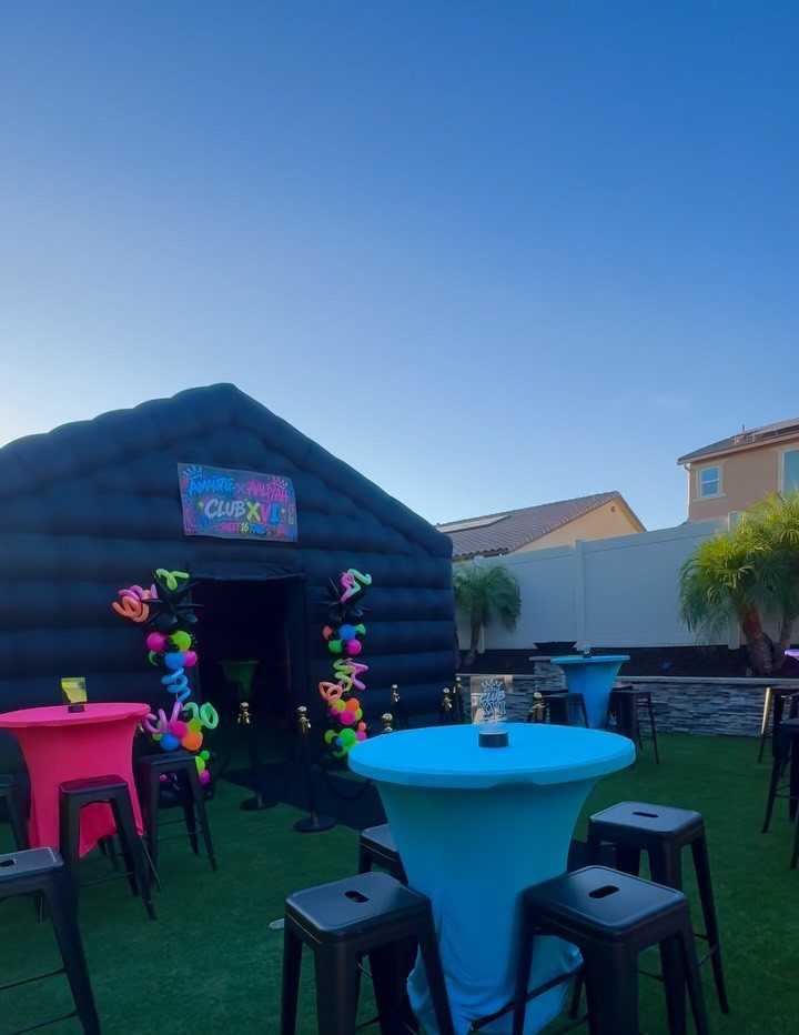 Outdoor party setup with a black inflatable club, colorful tables, and balloon arches.