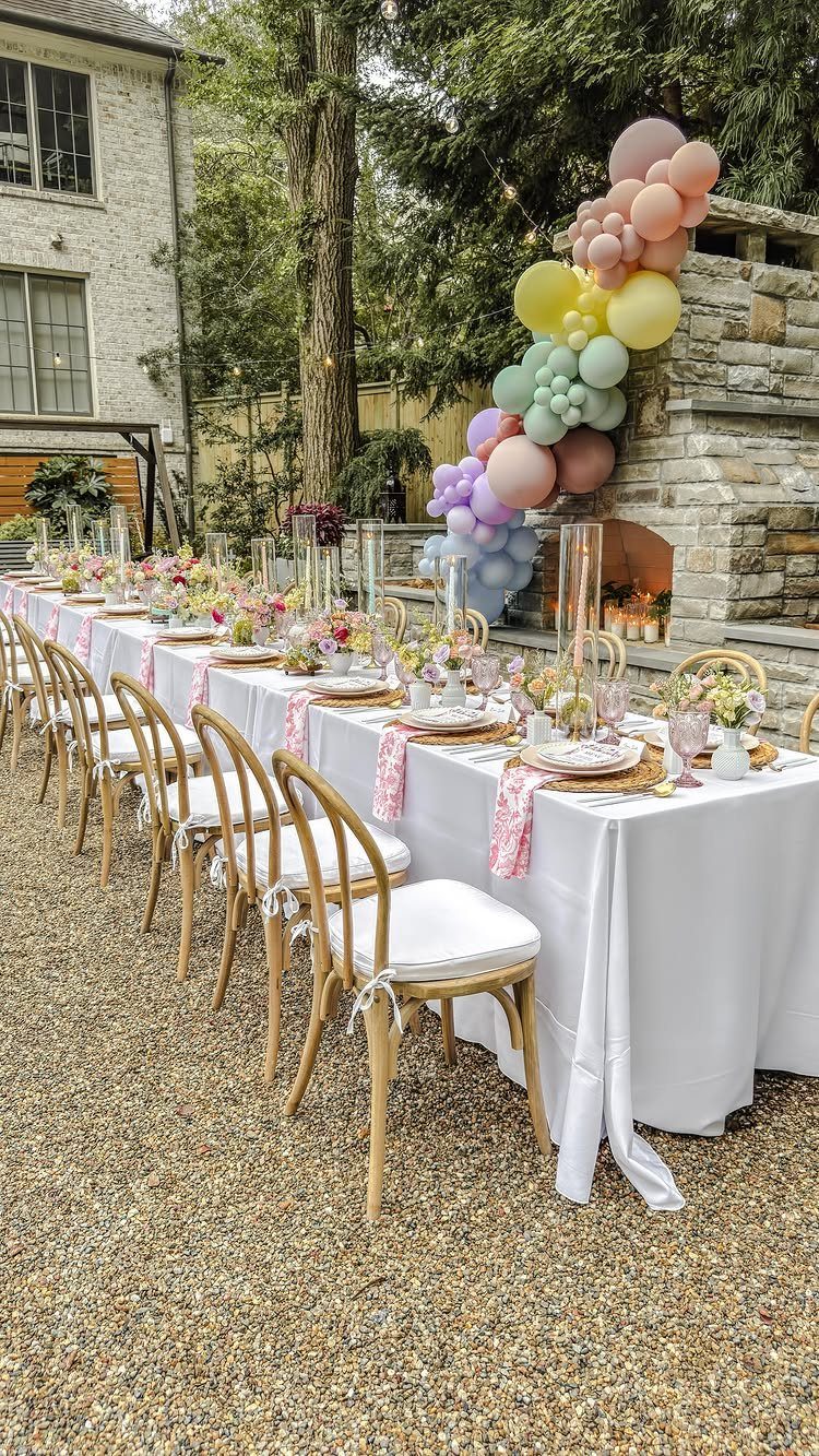 Outdoor party table with white linens, gold chairs, and pastel balloon arch.