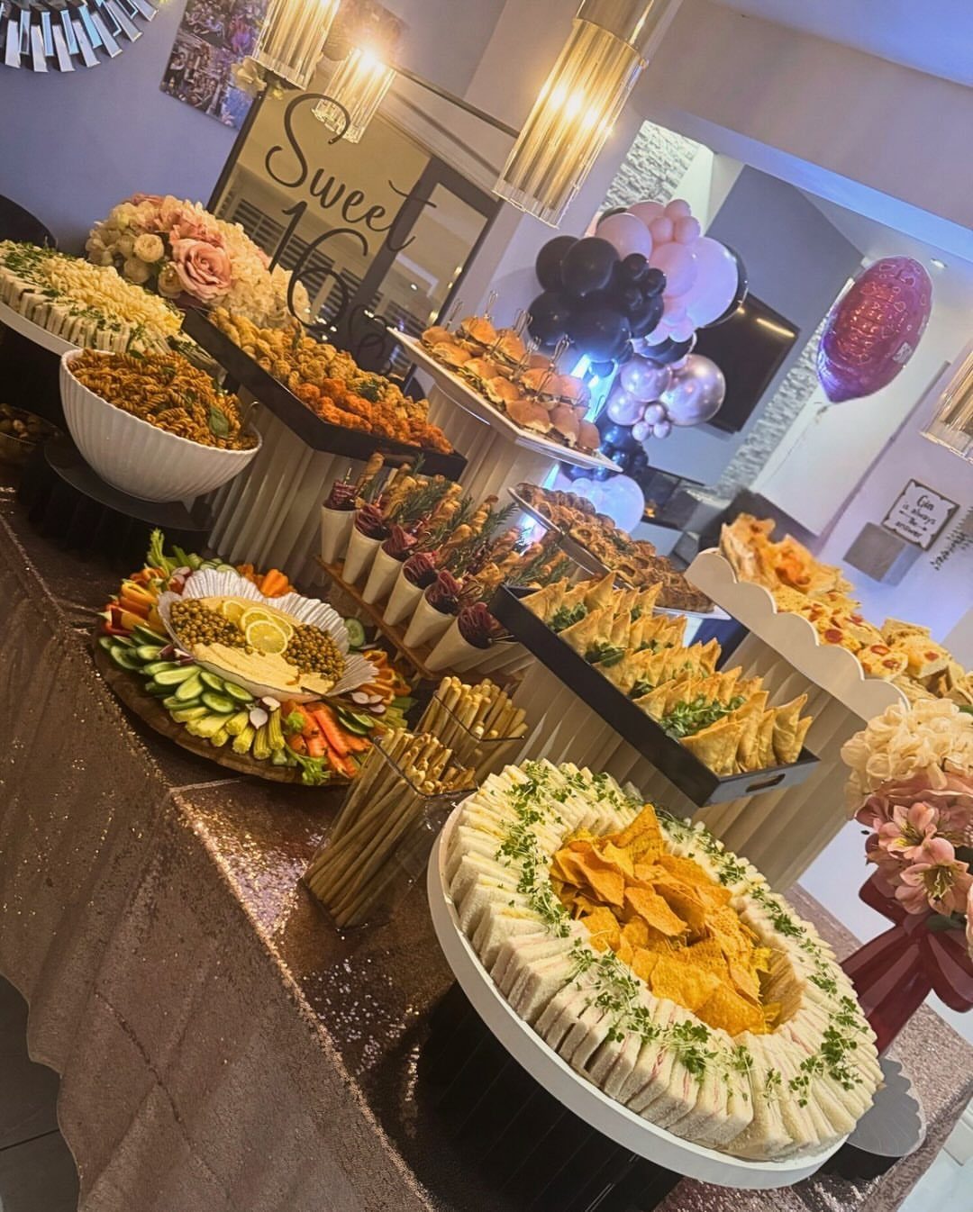Party food spread on gold sequin table with Sweet 16 sign and balloons.