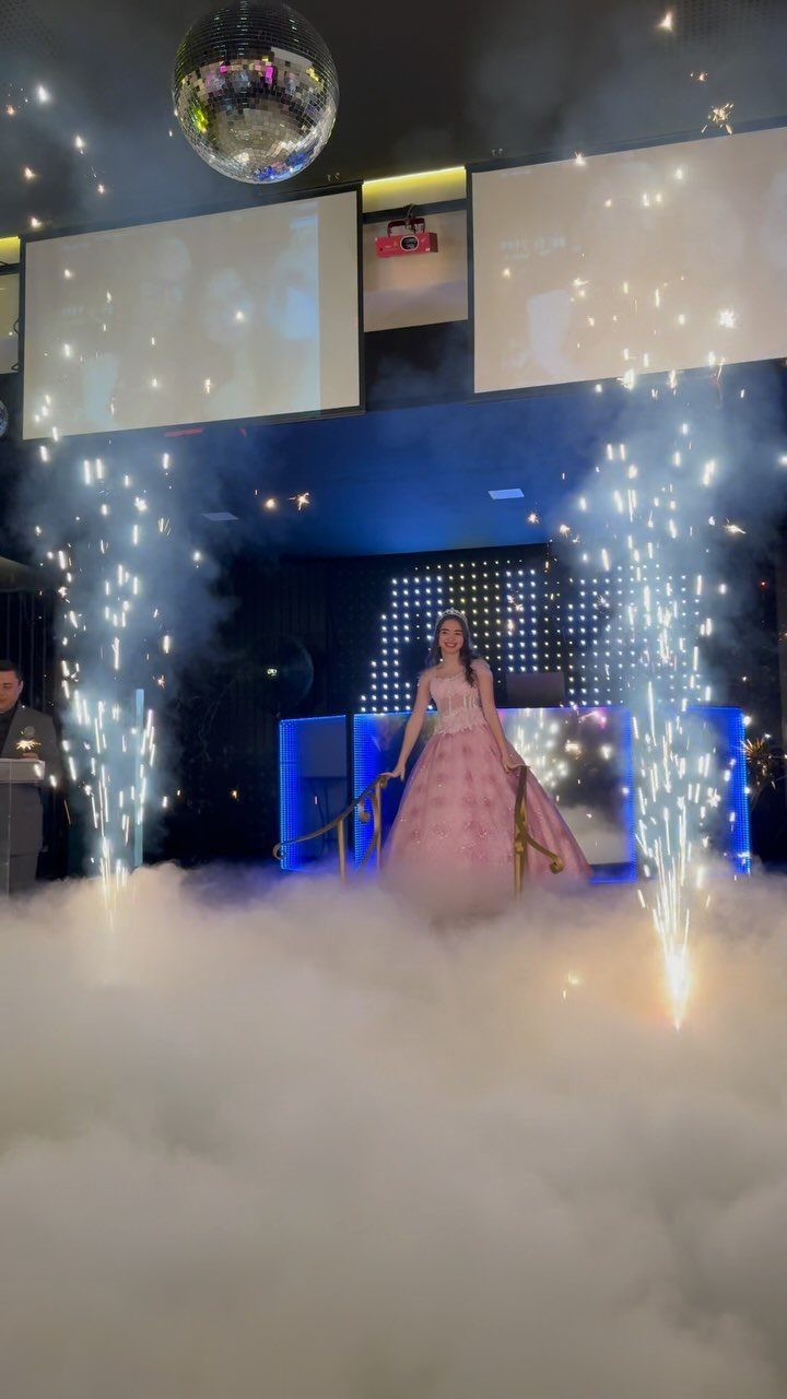 Young woman in pink gown descending stairs with fog and pyrotechnic sparks.