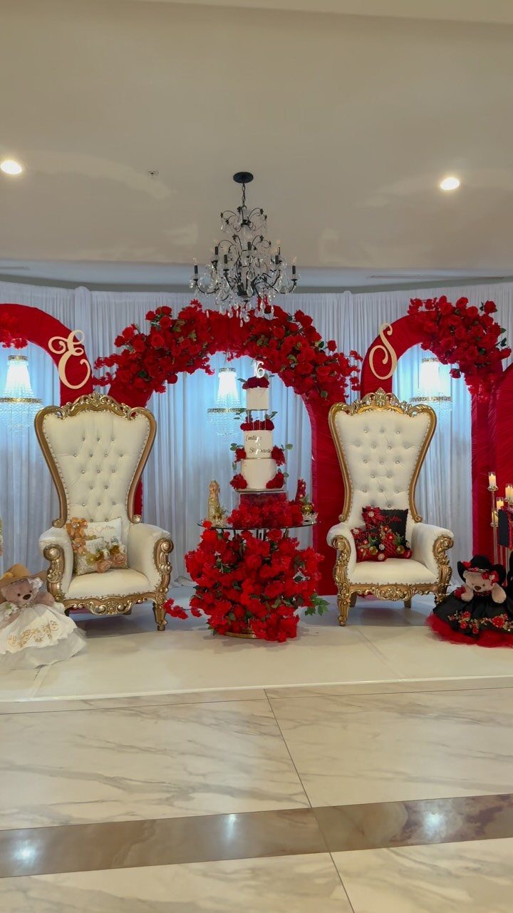 Ornate white chairs and a tiered cake surrounded by red roses and white drapery.