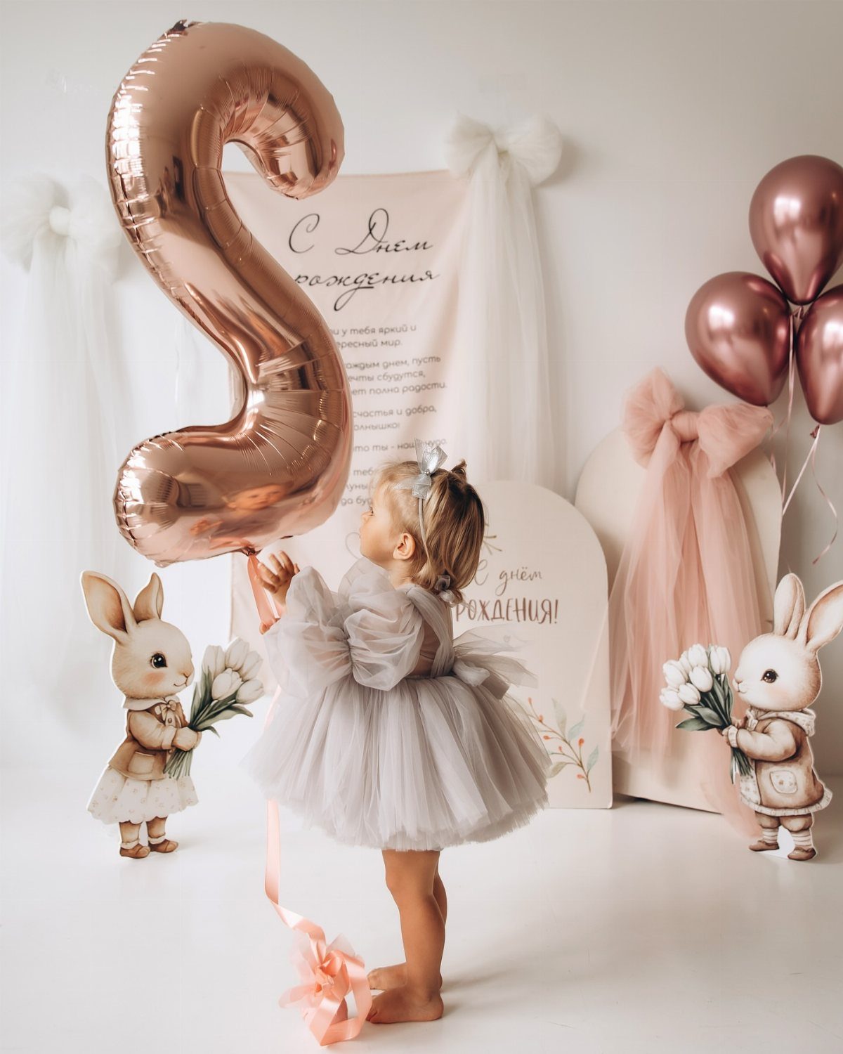 Young girl in grey tulle dress posing with rose-gold balloons and rabbit figurines.