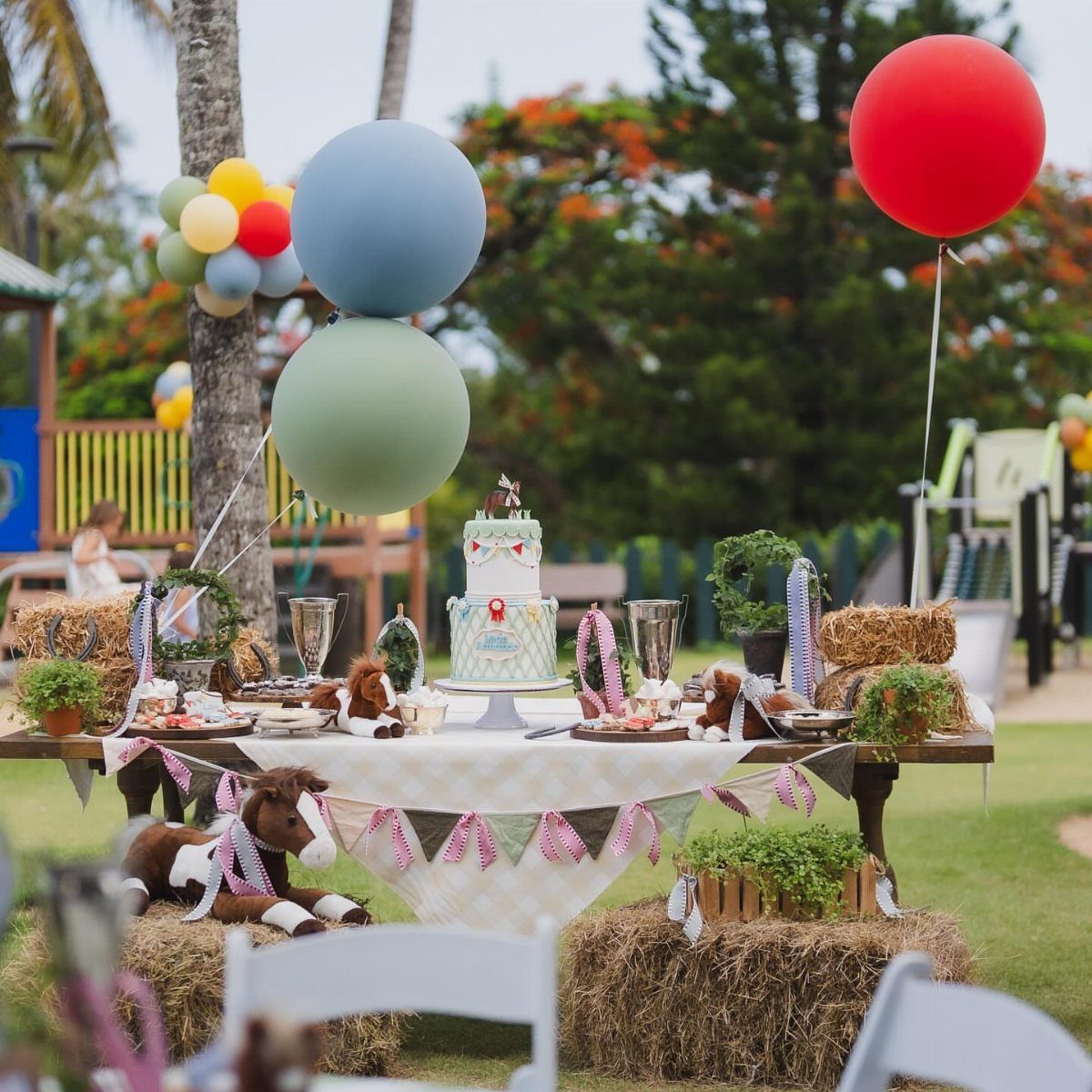 Party table with cake, balloons, and horse figurines set against hay bales outdoors.