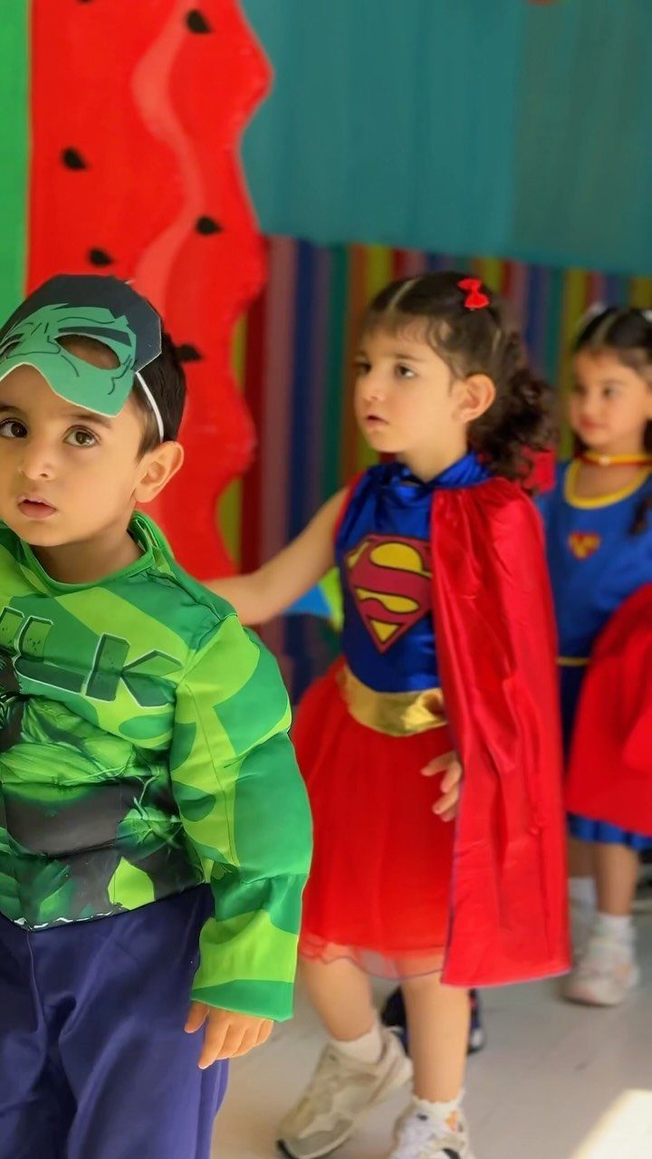 Three toddlers wearing superhero costumes standing together in a colorful indoor setting.