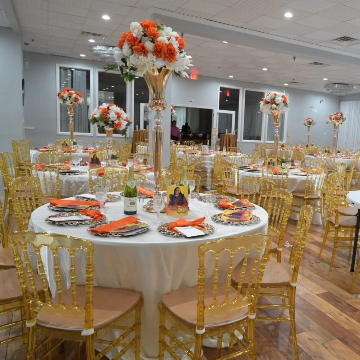 Banquet hall with white tablecloths, gold chairs, and orange floral centerpieces for a party.