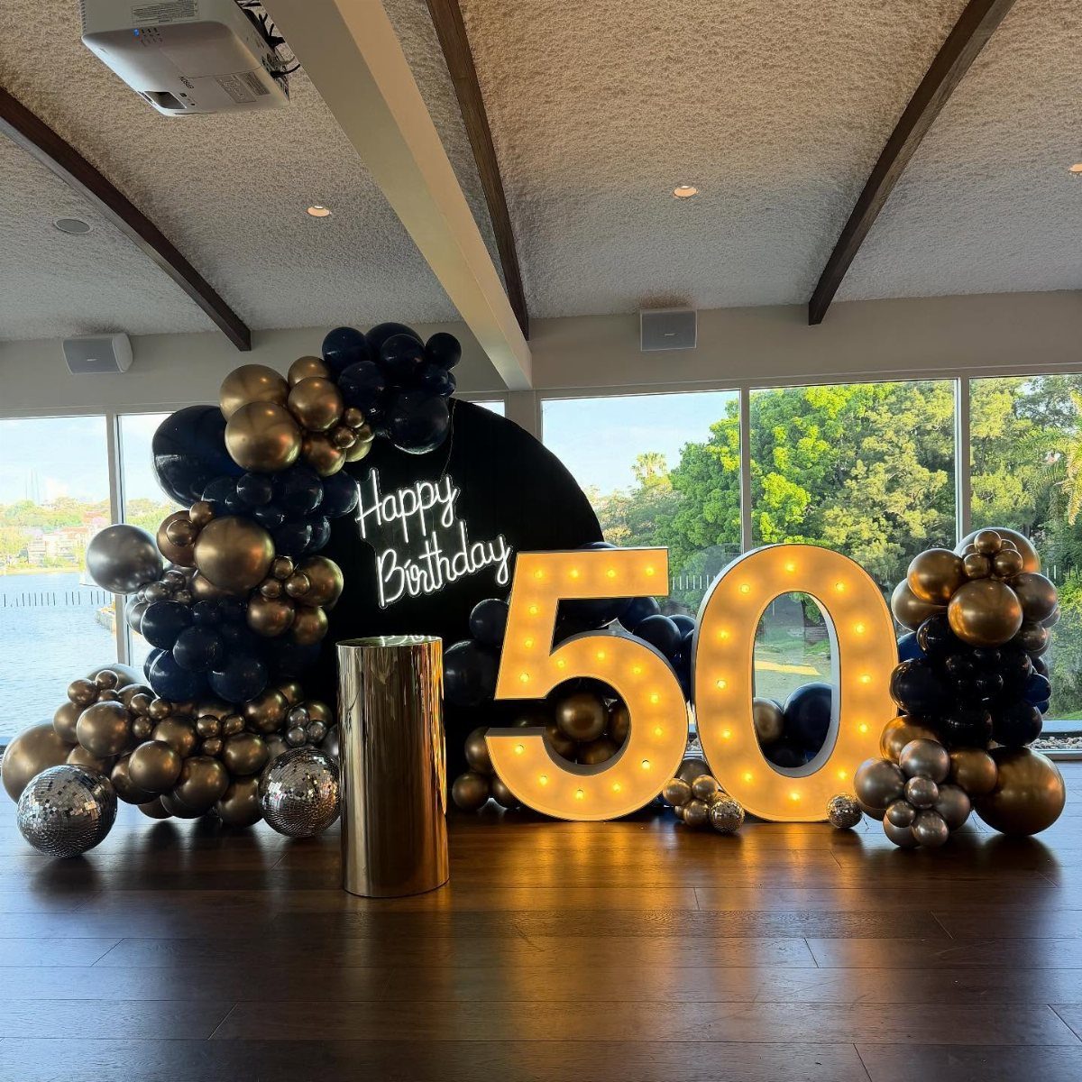 Glowing gold number 50 lights surrounded by black and gold balloons and backdrop.