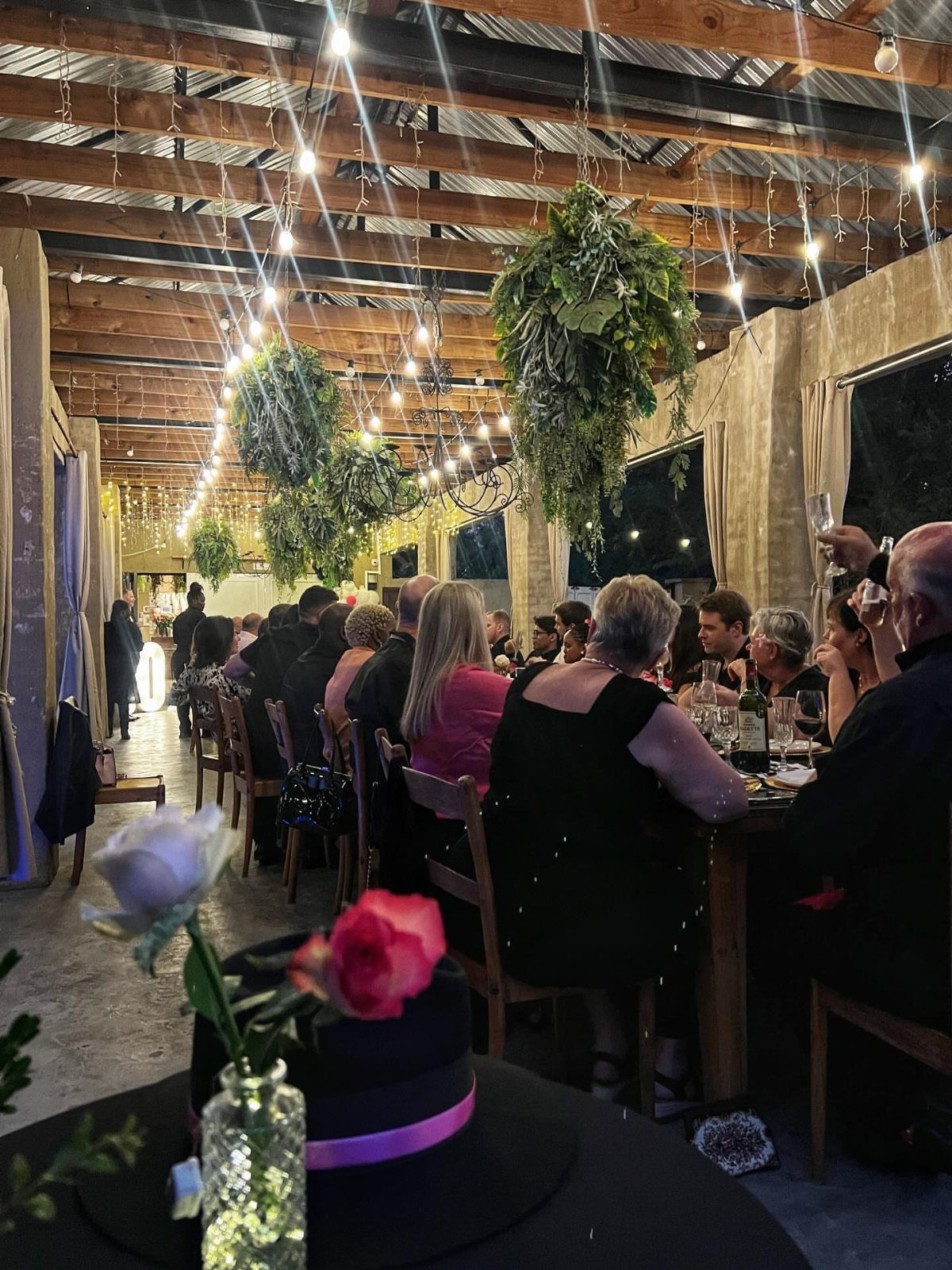 Rustic event venue with string lights, hanging plants, and guests dining at tables.