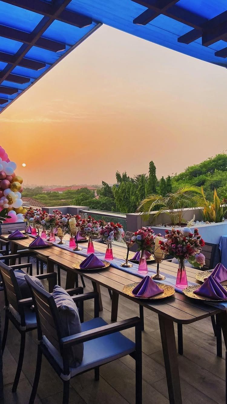 Rooftop dining table set with purple napkins and flowers overlooking a sunset landscape.