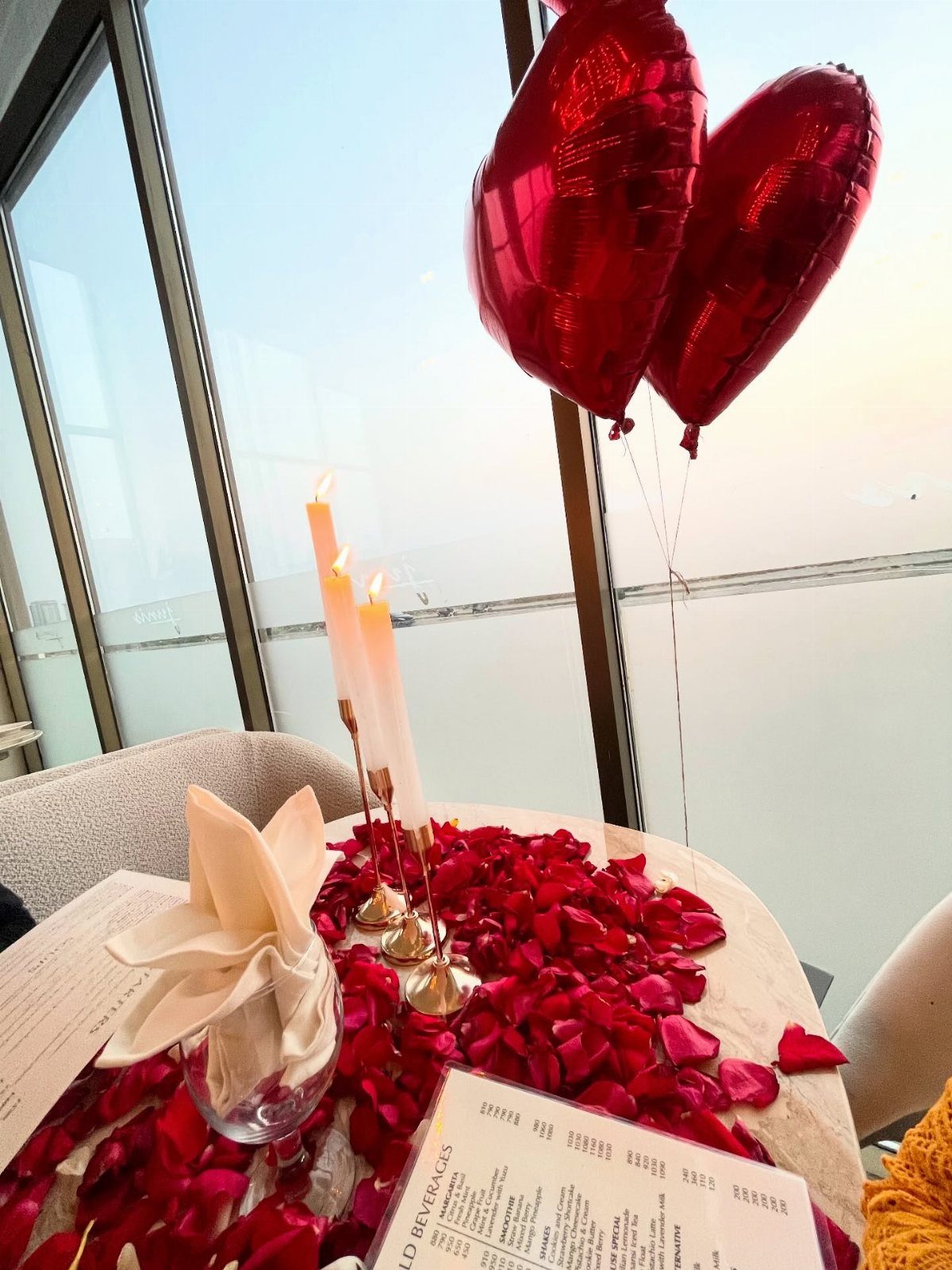 Romantic table setting with red rose petals, candles, and heart balloons by a window.
